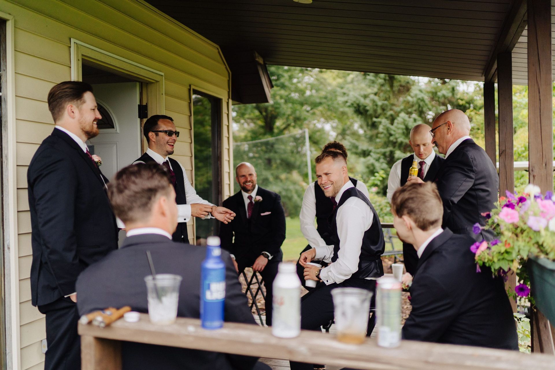 Groomsmen on a porch: men in suits and vests, some holding drinks and cigars, laughing and socializing.