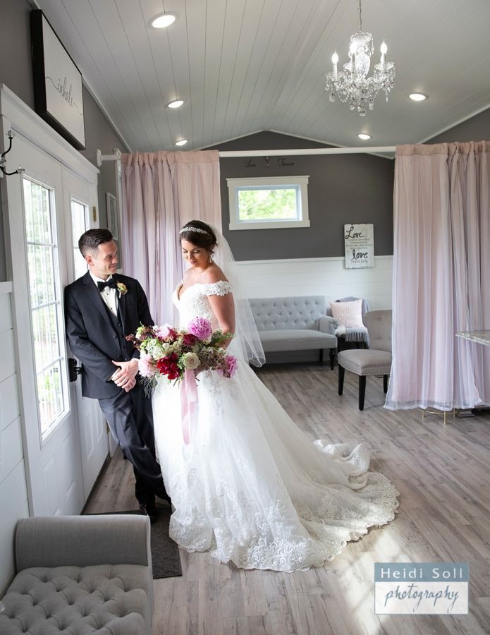 Bride and groom in elegant bridal suite. Bride holds bouquet, groom gazes at her near window. Pastel decor, chandelier.