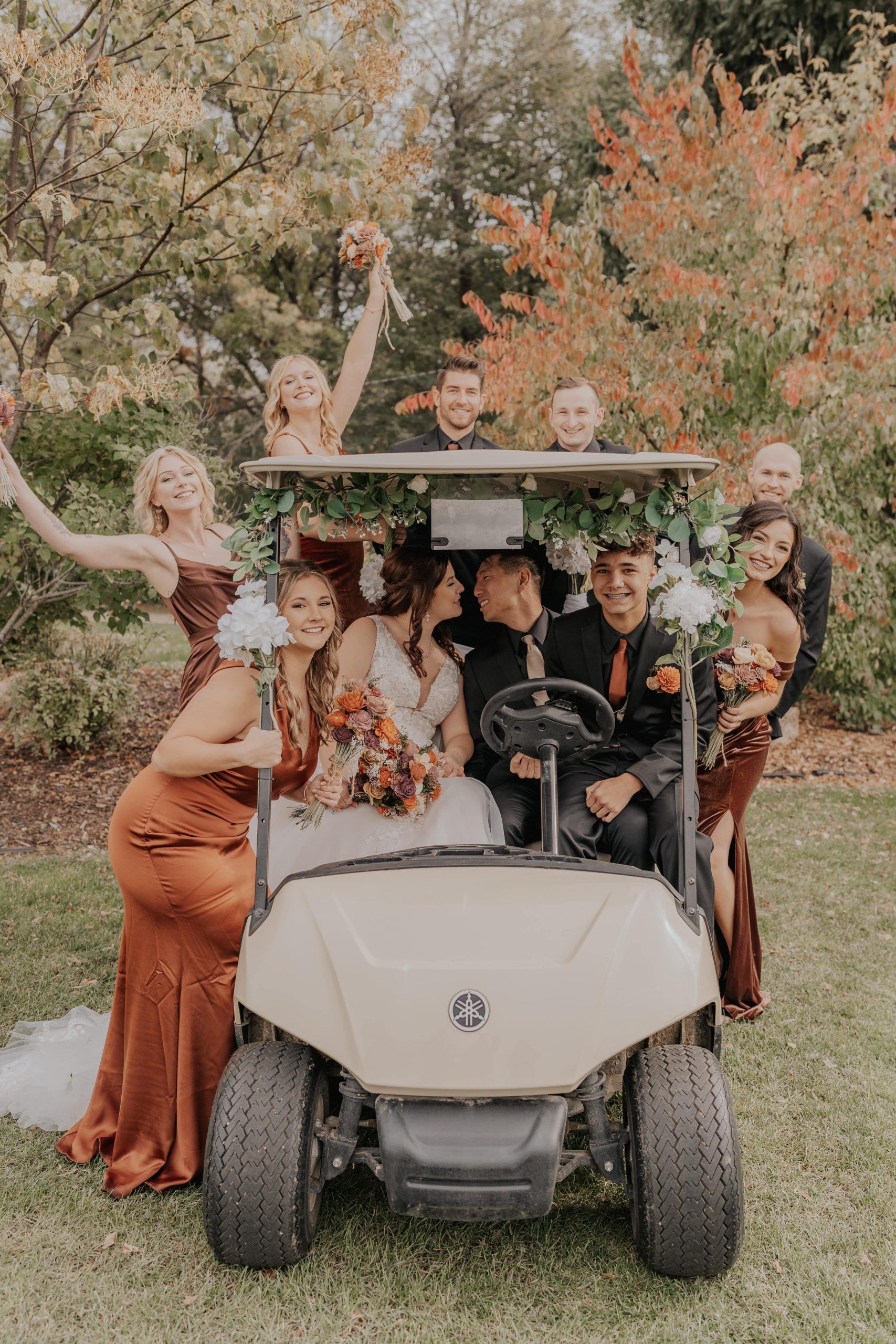 Wedding party in a golf cart; bride and groom kiss, others cheer. Autumn colors, outdoor setting.
