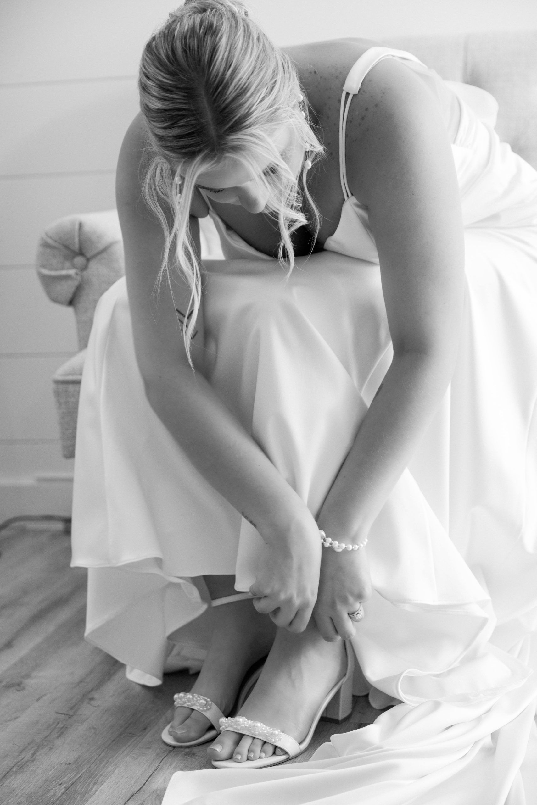 Bride seated, adjusting her shoe. Wearing a white wedding gown, pearl bracelet, in a soft-lit room.