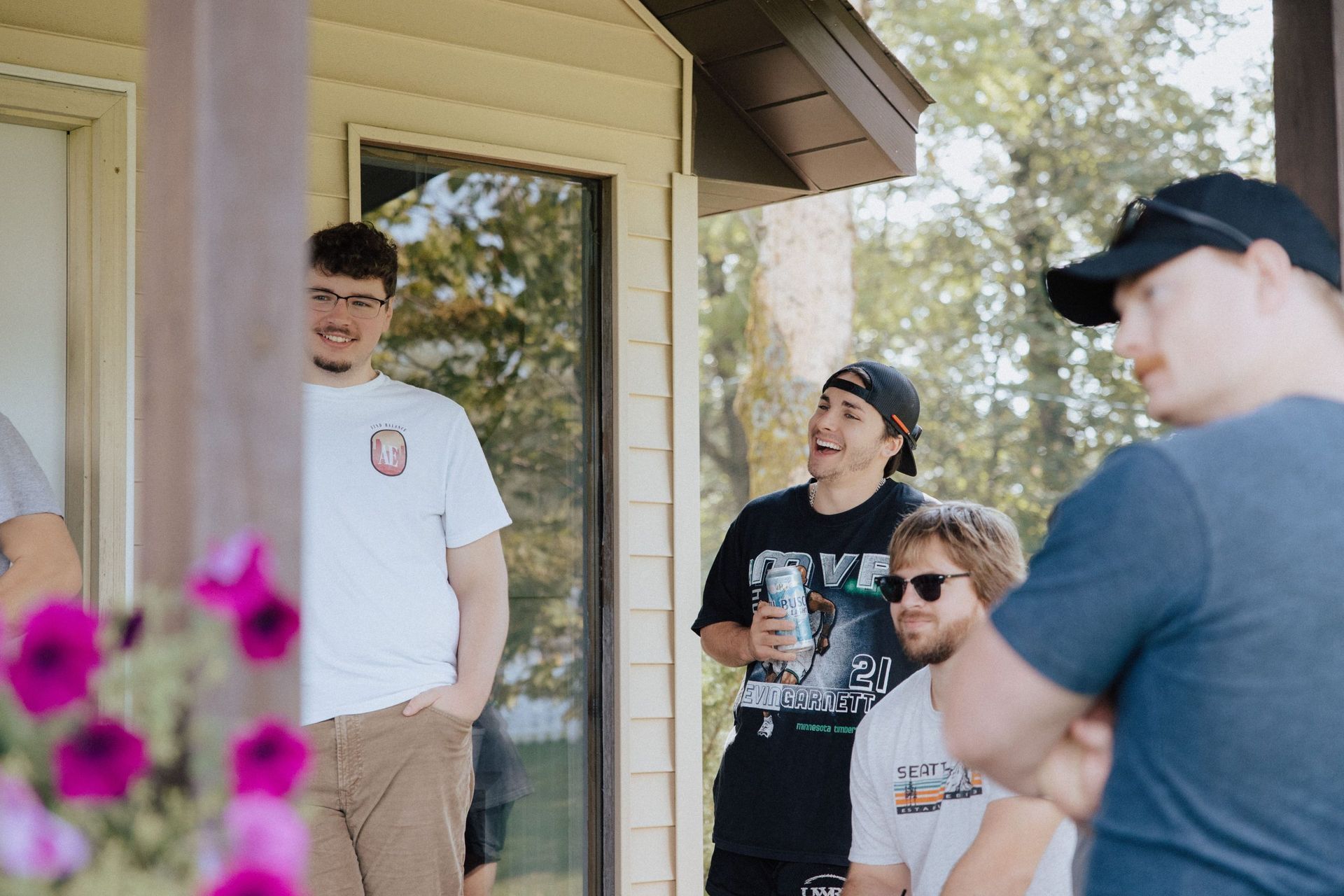 Group of young men on a porch, smiling and talking. One man holds a can. Sunny day.