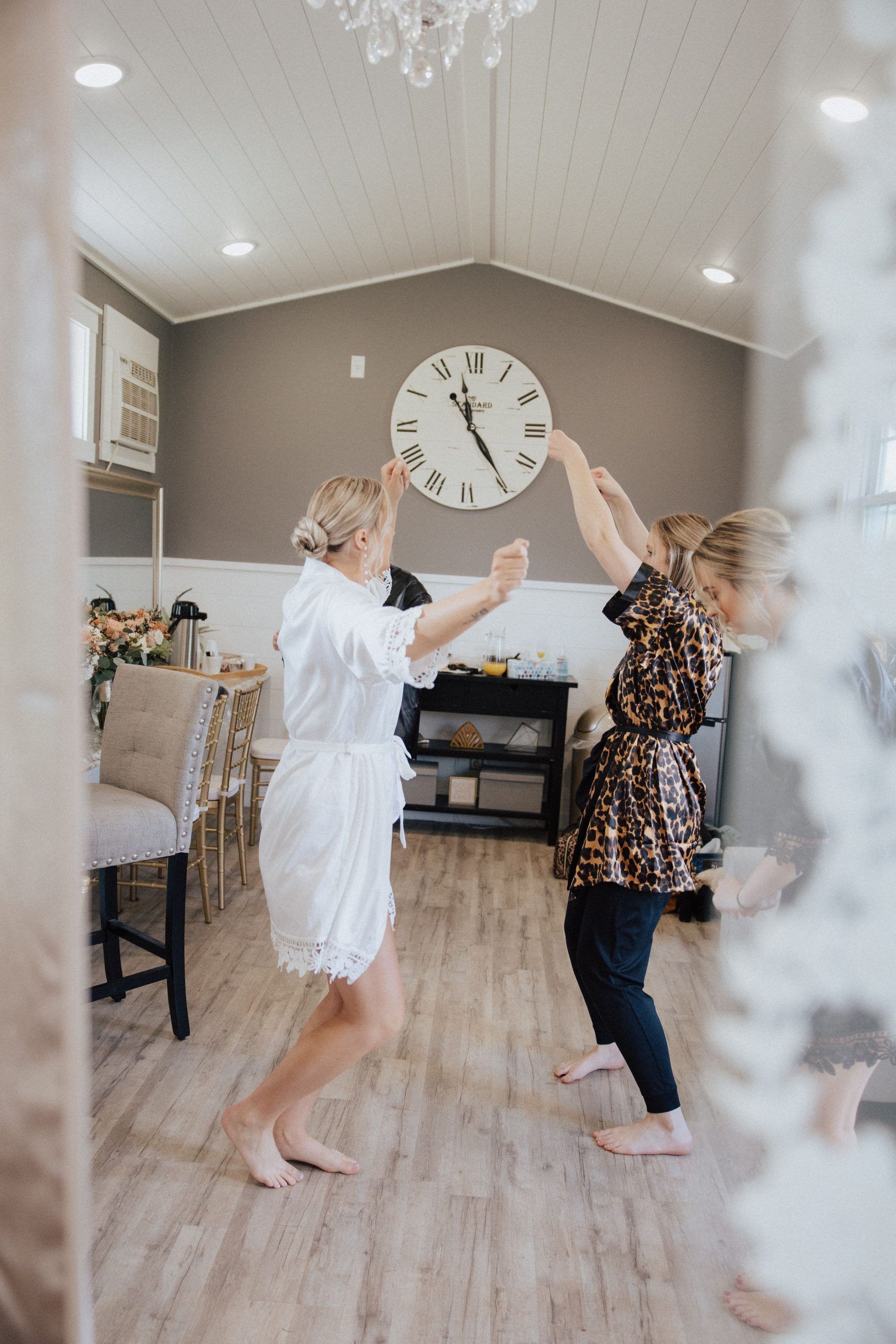 Two barefoot women dancing in a room; one in a white robe, the other in animal print; a clock on the wall.