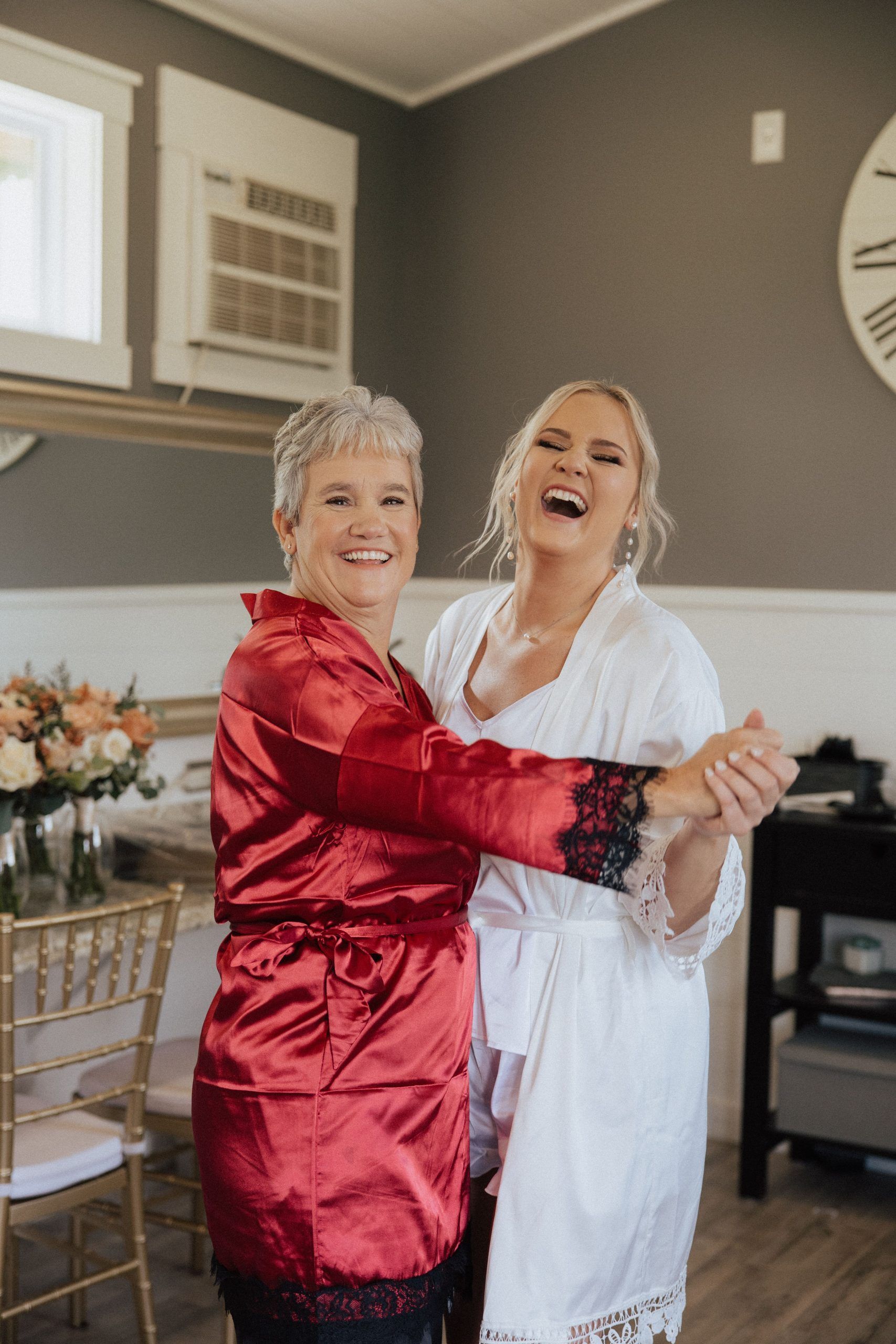Mother and daughter laughing, dancing together in a room. Mother wears red robe. Daughter wears white robe.