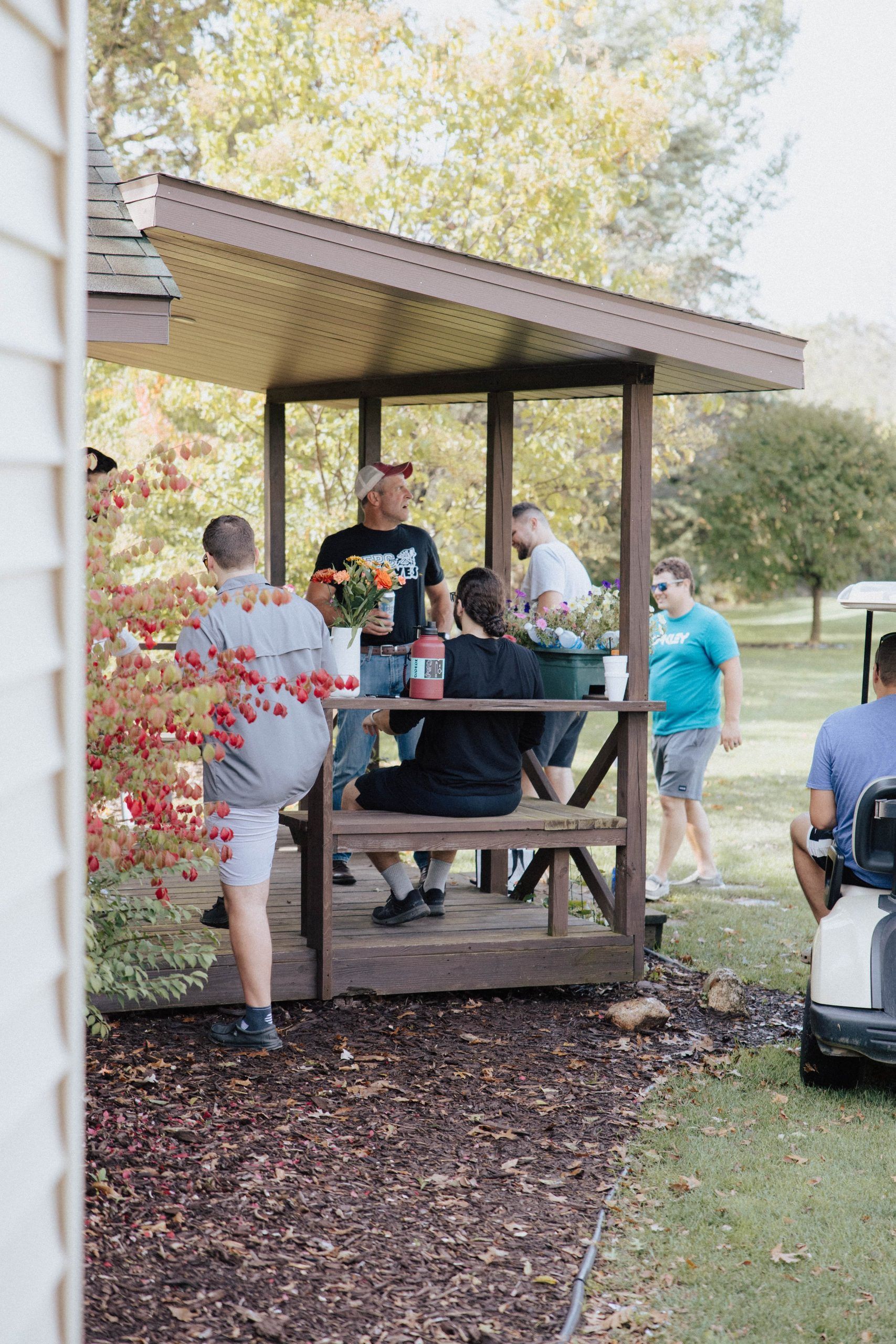 People gather around a brown gazebo outdoors; some holding flowers and conversing.