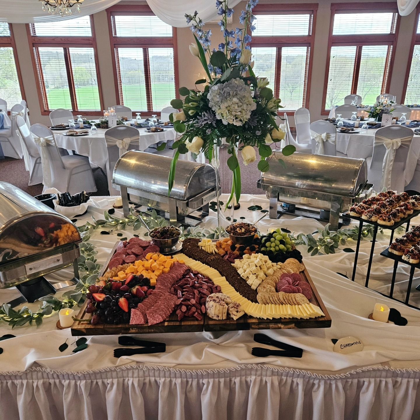 Buffet table with charcuterie board, flowers, and chafing dishes at a wedding reception with white linens.