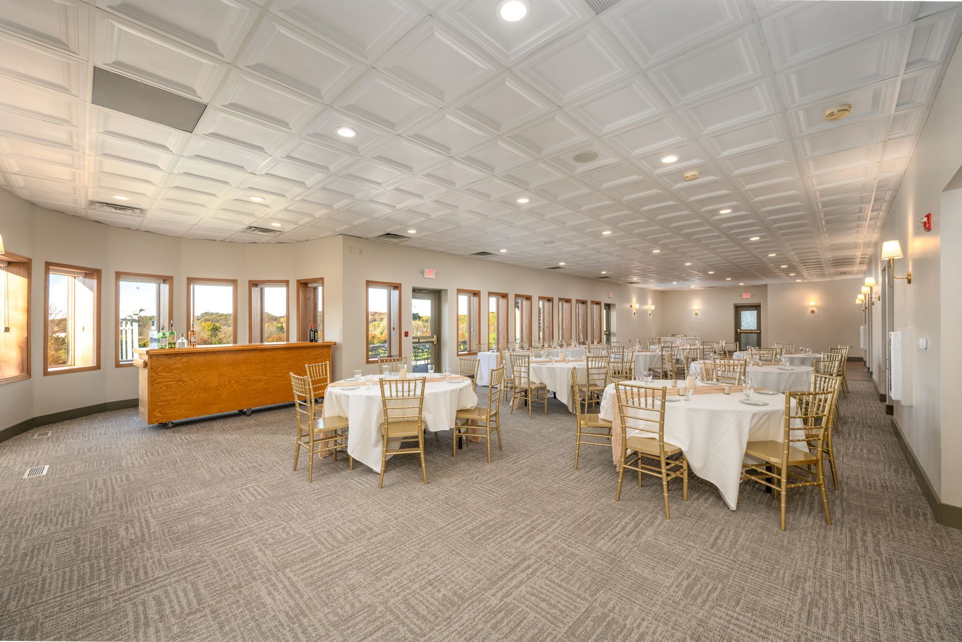 Empty event space with round tables set for a gathering, cream-colored chairs, and a bar area.