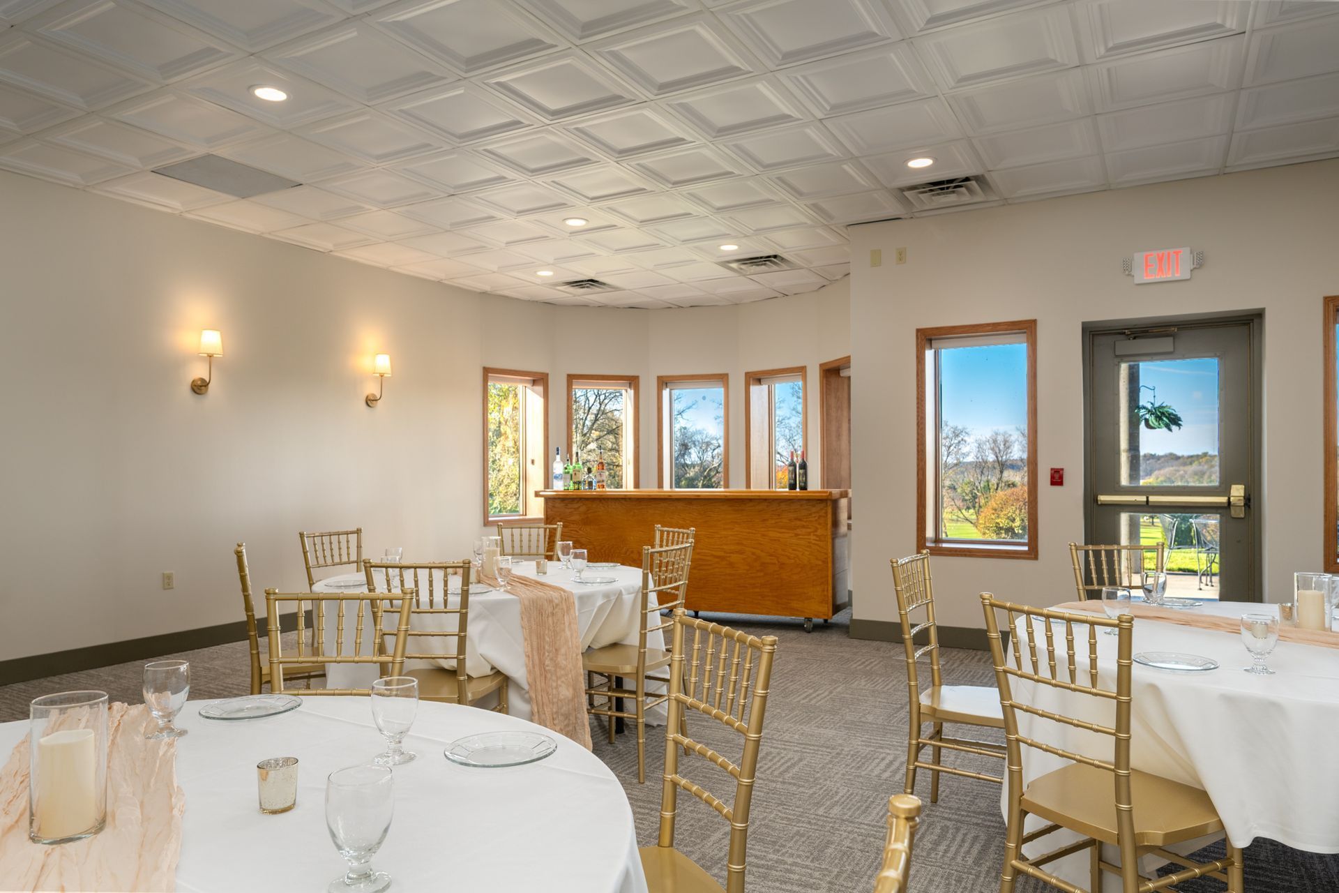 A banquet hall with round tables set for a meal.  Gold chairs and a bar are visible. Windows offer a view.