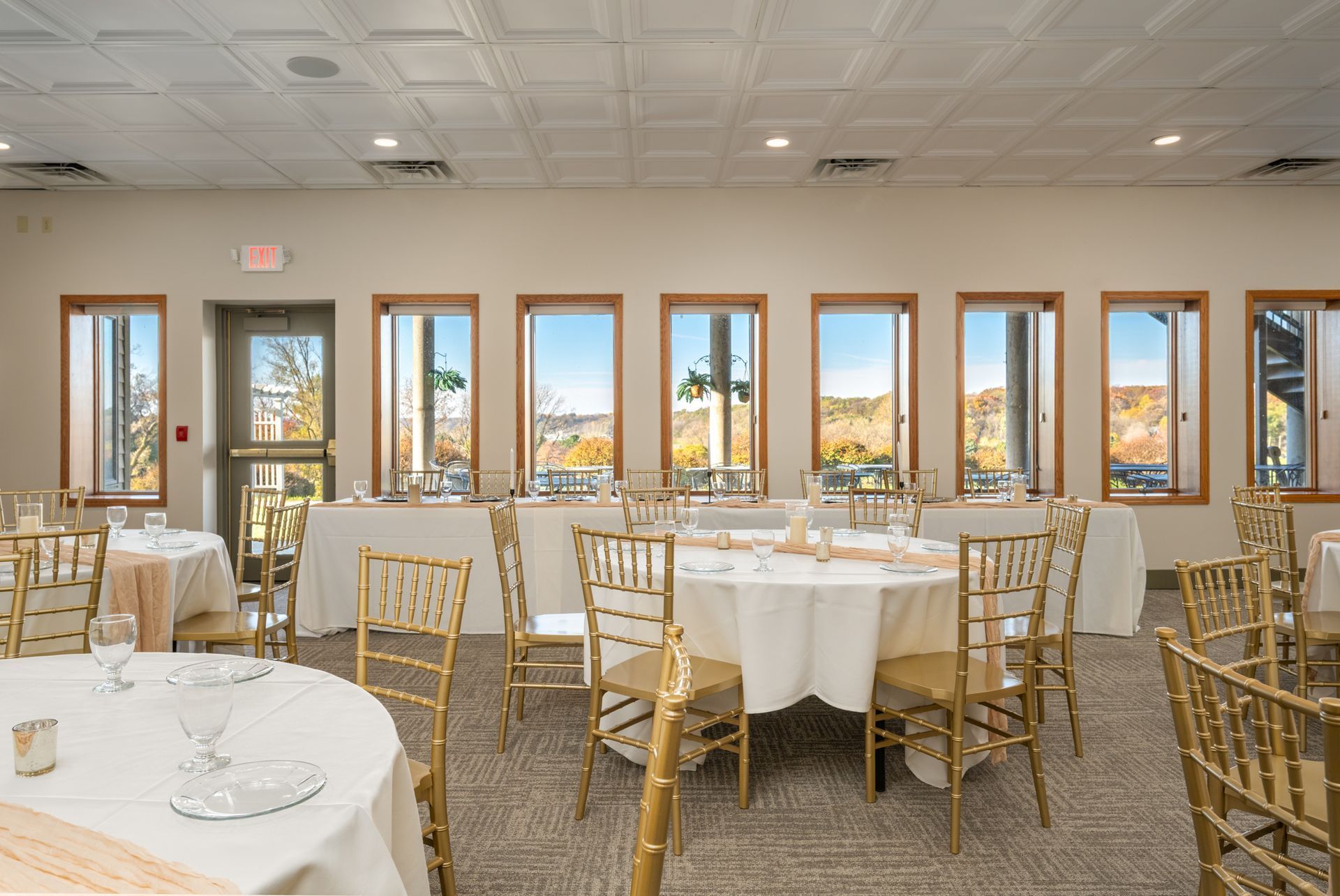 Banquet room with round tables, gold chairs, and windows overlooking a landscape.