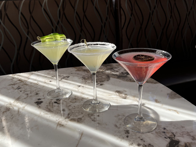 Three martini cocktails on a white table: green, clear, and pink, each with a garnish.