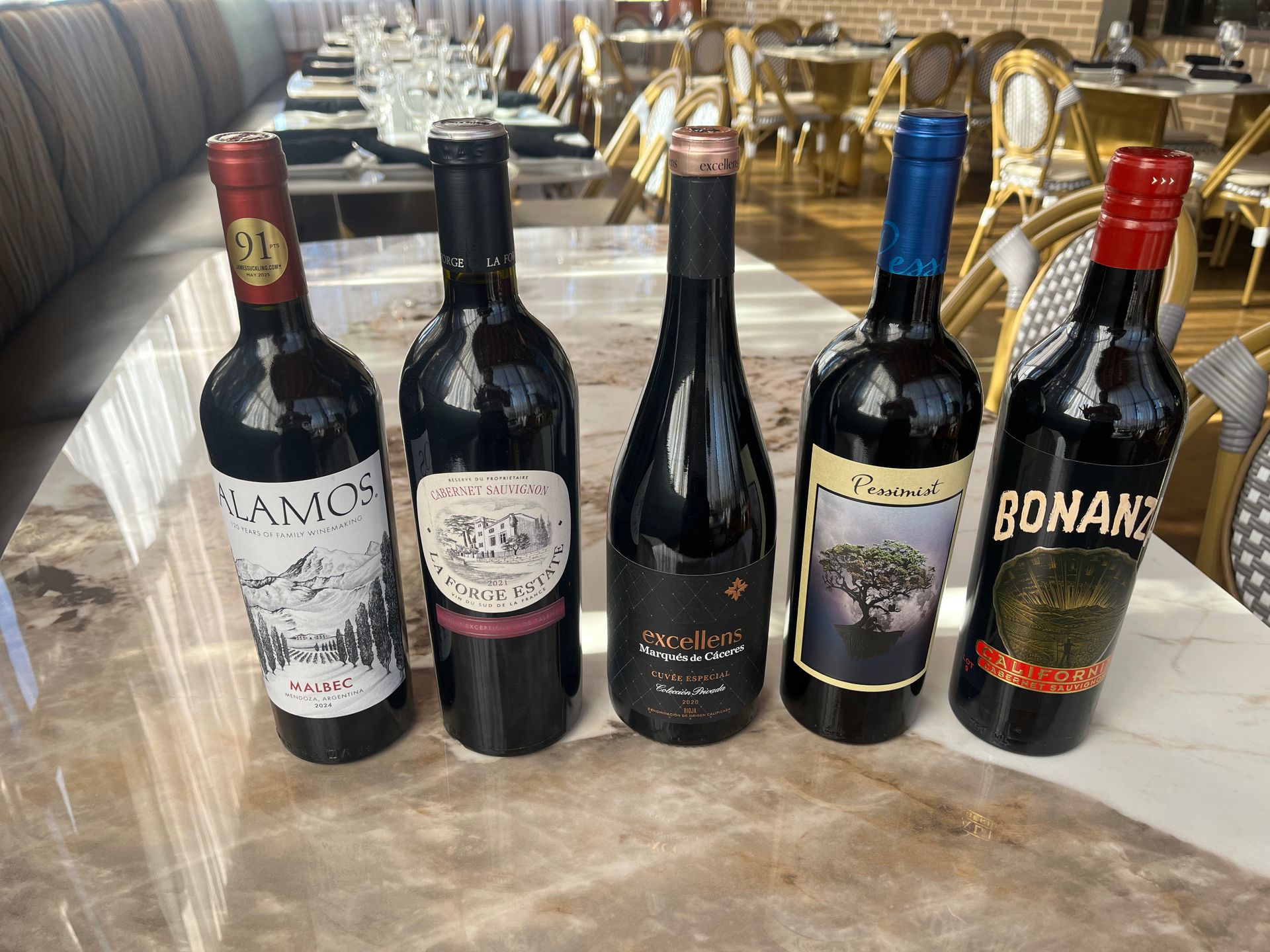 Five red wine bottles on a marble table at an outdoor restaurant.