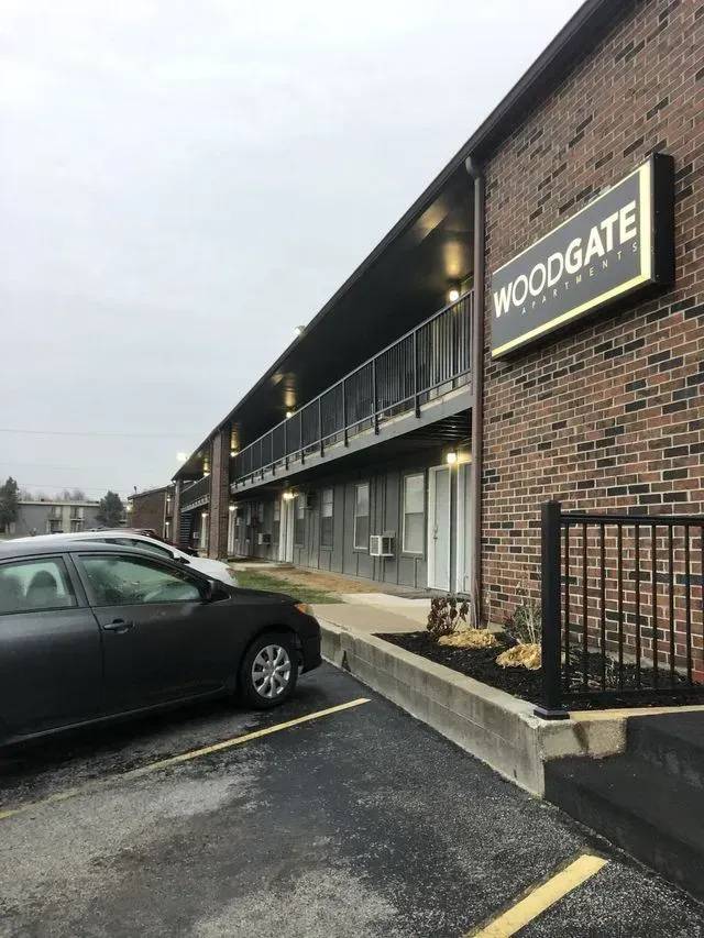 Woodgate Apartments building exterior, brick, with a black and gold sign, parking lot, overcast sky.