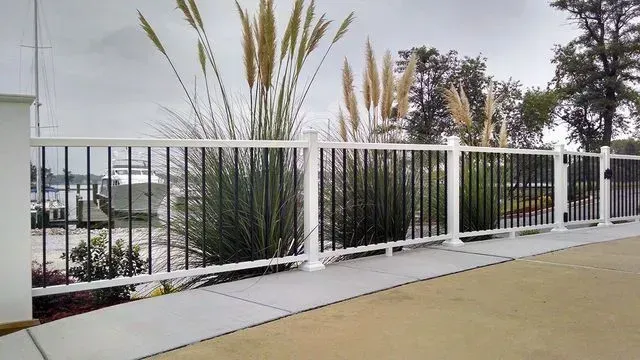 White and black metal fence with tall ornamental grasses and water visible in the background.