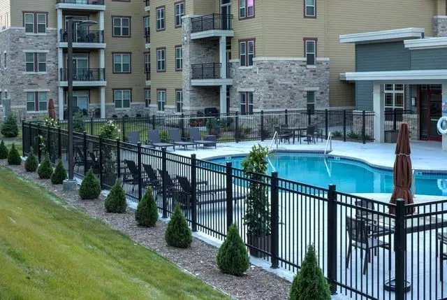 Apartment building exterior with a pool, black fence, and small trees on a grassy hill.