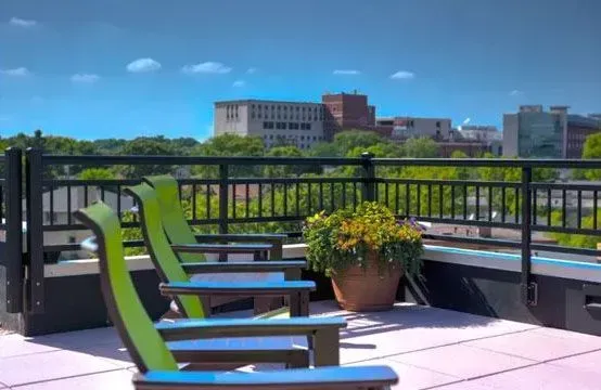 Rooftop patio with green chairs, potted flowers, and a cityscape view under a bright blue sky.