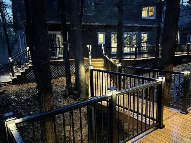 Dark wooden deck and stairs with illuminated railings, leading to a house with lit windows, surrounded by trees.
