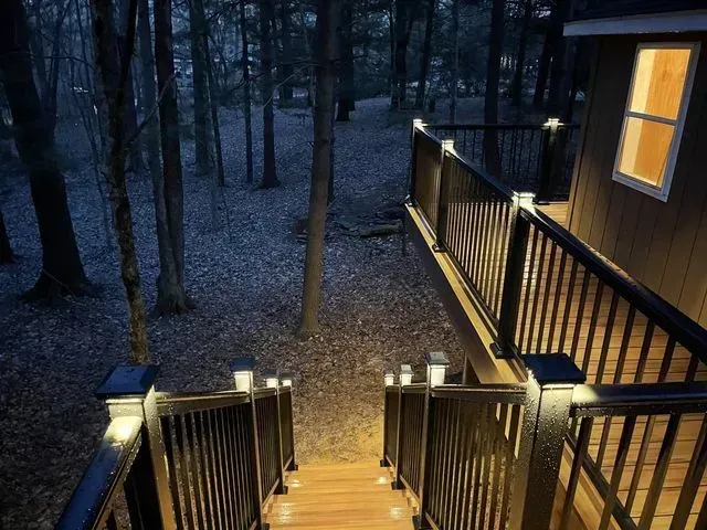 Wooden deck and stairs with lights leading into a dark forest, lit window on the right.