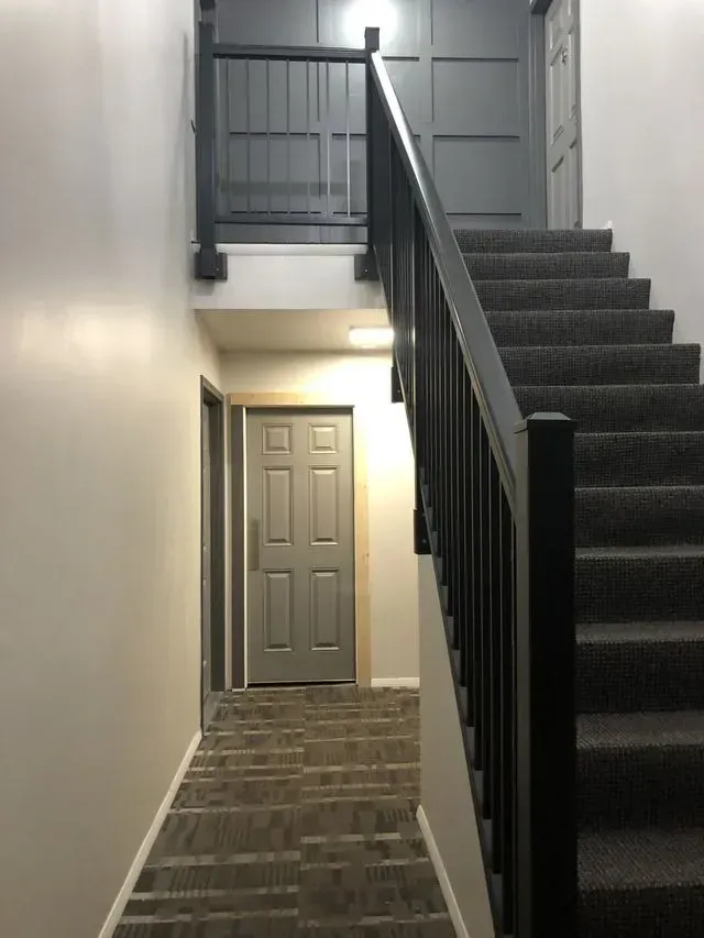 Stairwell with carpeted stairs, gray walls, and a black handrail leading to a closed gray door.