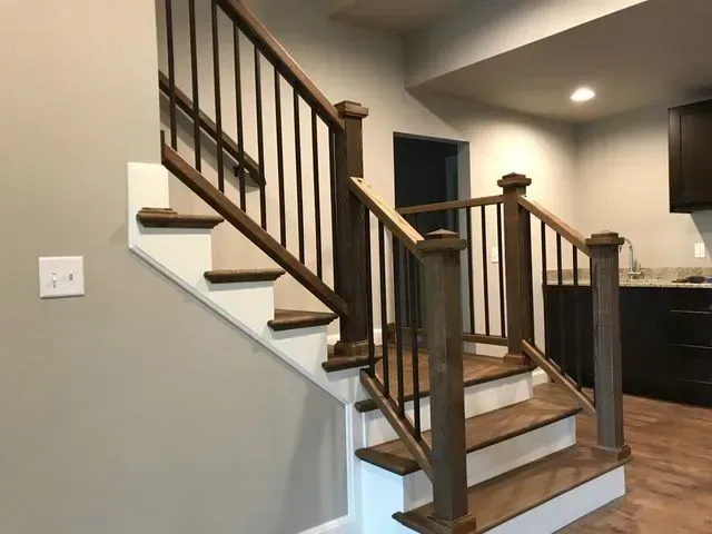 Staircase with wooden handrails and black metal balusters. White risers, brown treads, beige walls, and dark cabinets visible.