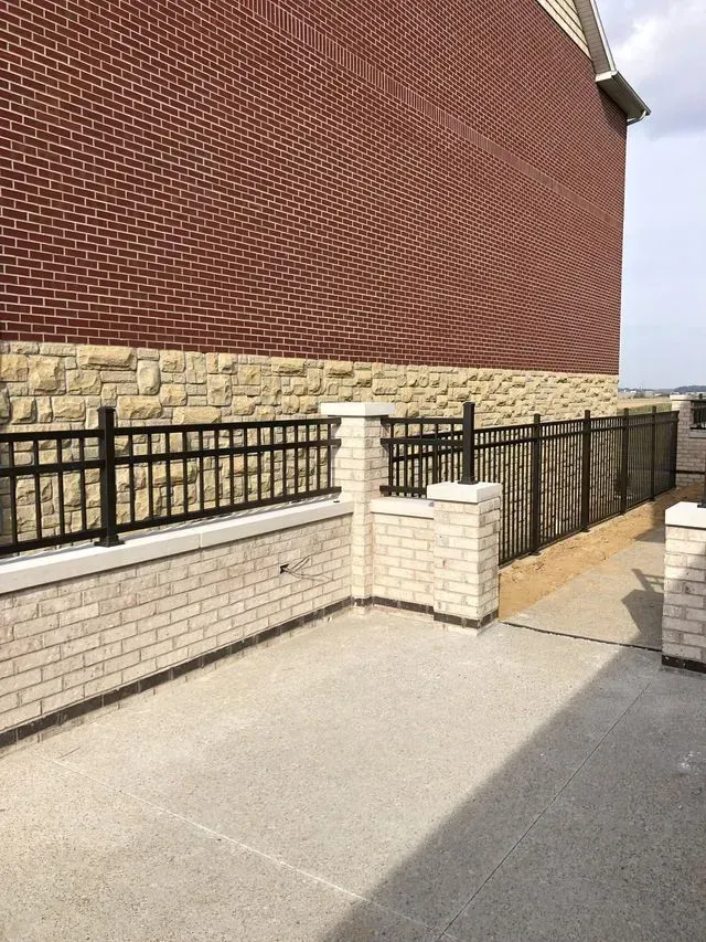 Brick wall with a black metal fence, beige brick and concrete surface, under an overcast sky.