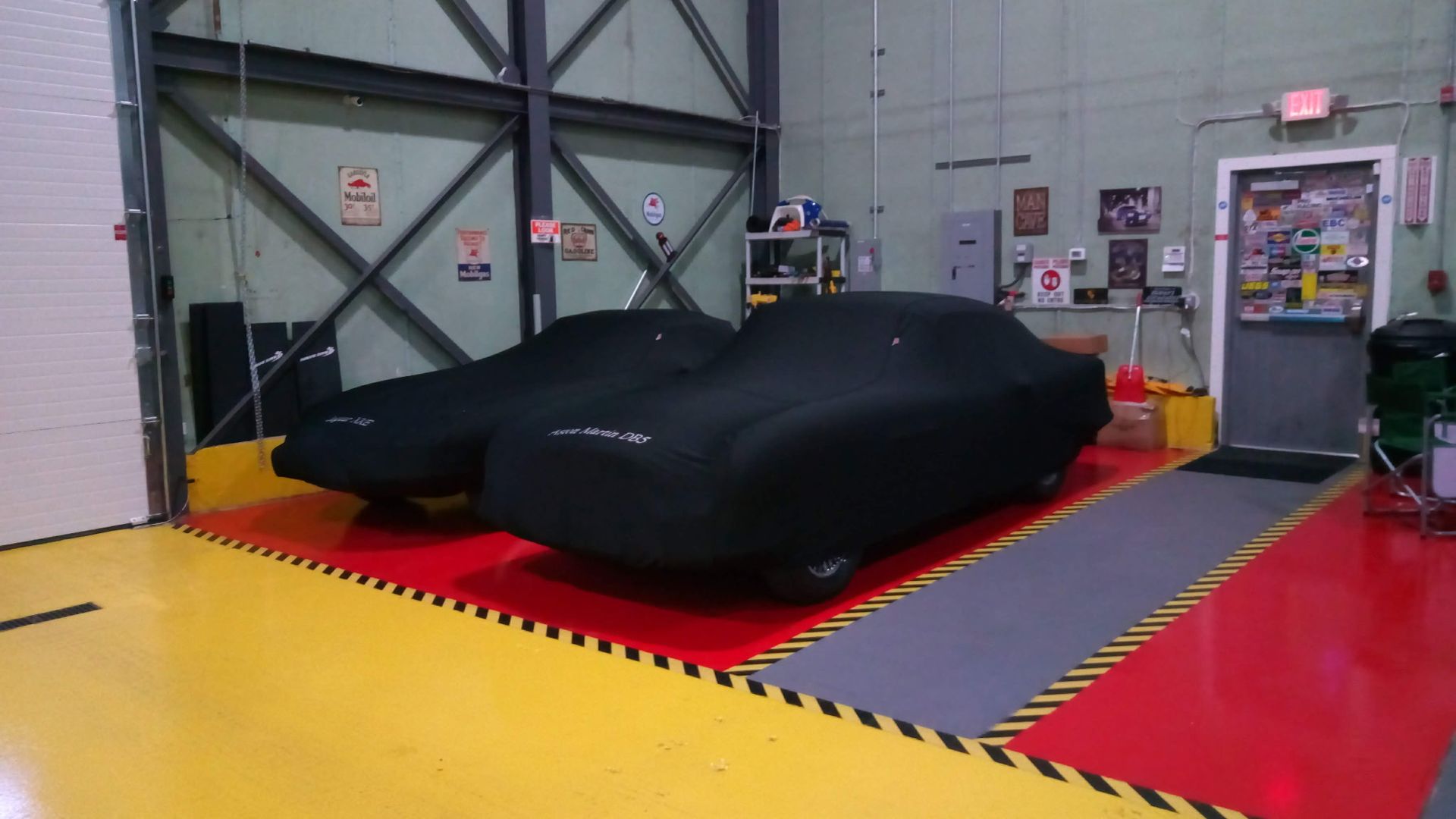 Black car under cover in an auto workshop on a red platform, with yellow floor and garage doors.