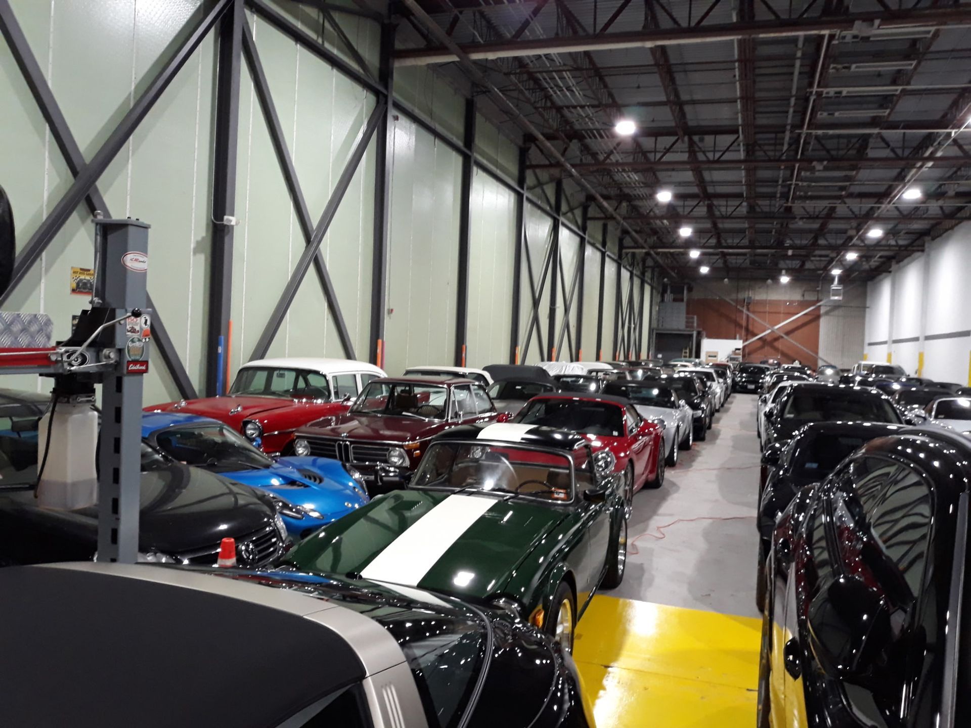 Rows of cars parked inside a large warehouse, including a black car with a white racing stripe.