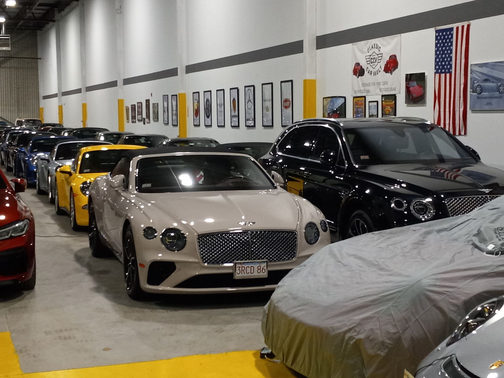 Indoor car showroom with several sports cars lined up, including a beige Bentley and covered vehicles.