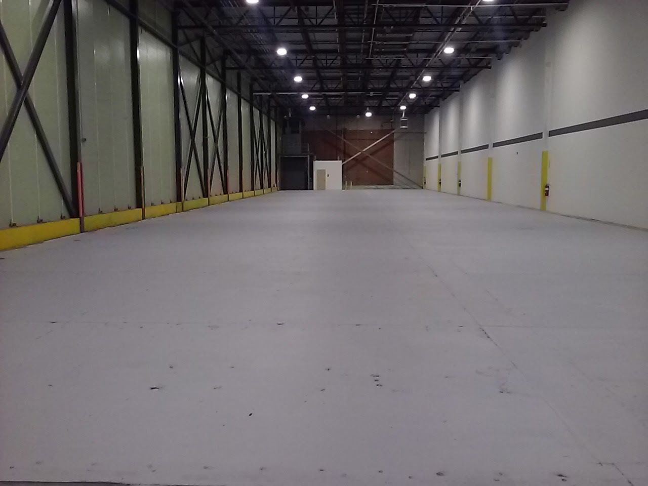 Empty industrial warehouse with a wide concrete floor and tall white walls under overhead lights