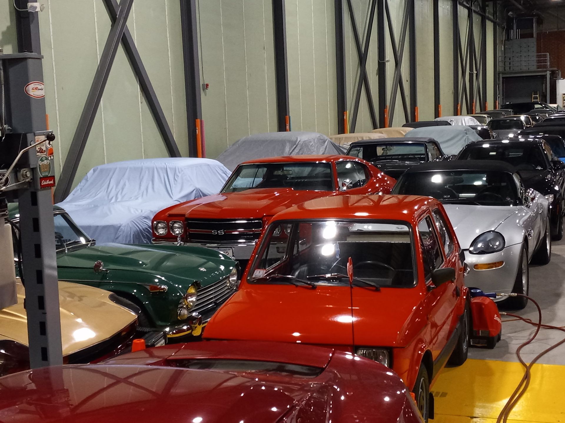 Classic cars parked indoors in a warehouse, including red, silver, green, and gold vehicles.