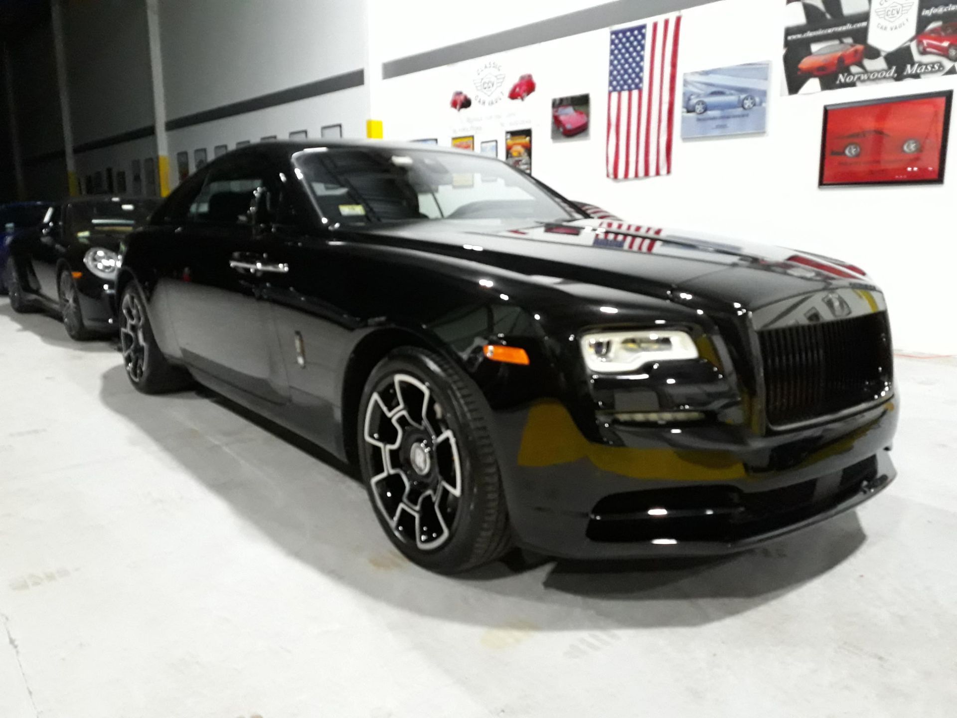 Black Rolls-Royce in a showroom, viewed from the front-left with an American flag on the wall behind it
