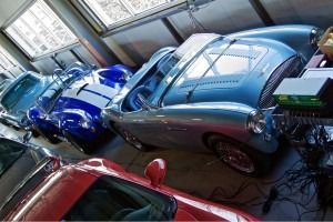 Three classic cars parked closely inside a garage, including blue, silver, and red cars.