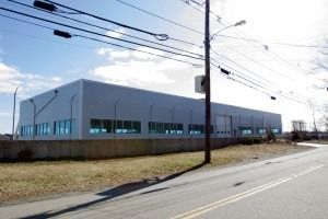 Large white industrial warehouse by a roadside under a bright sky, with power lines overhead.