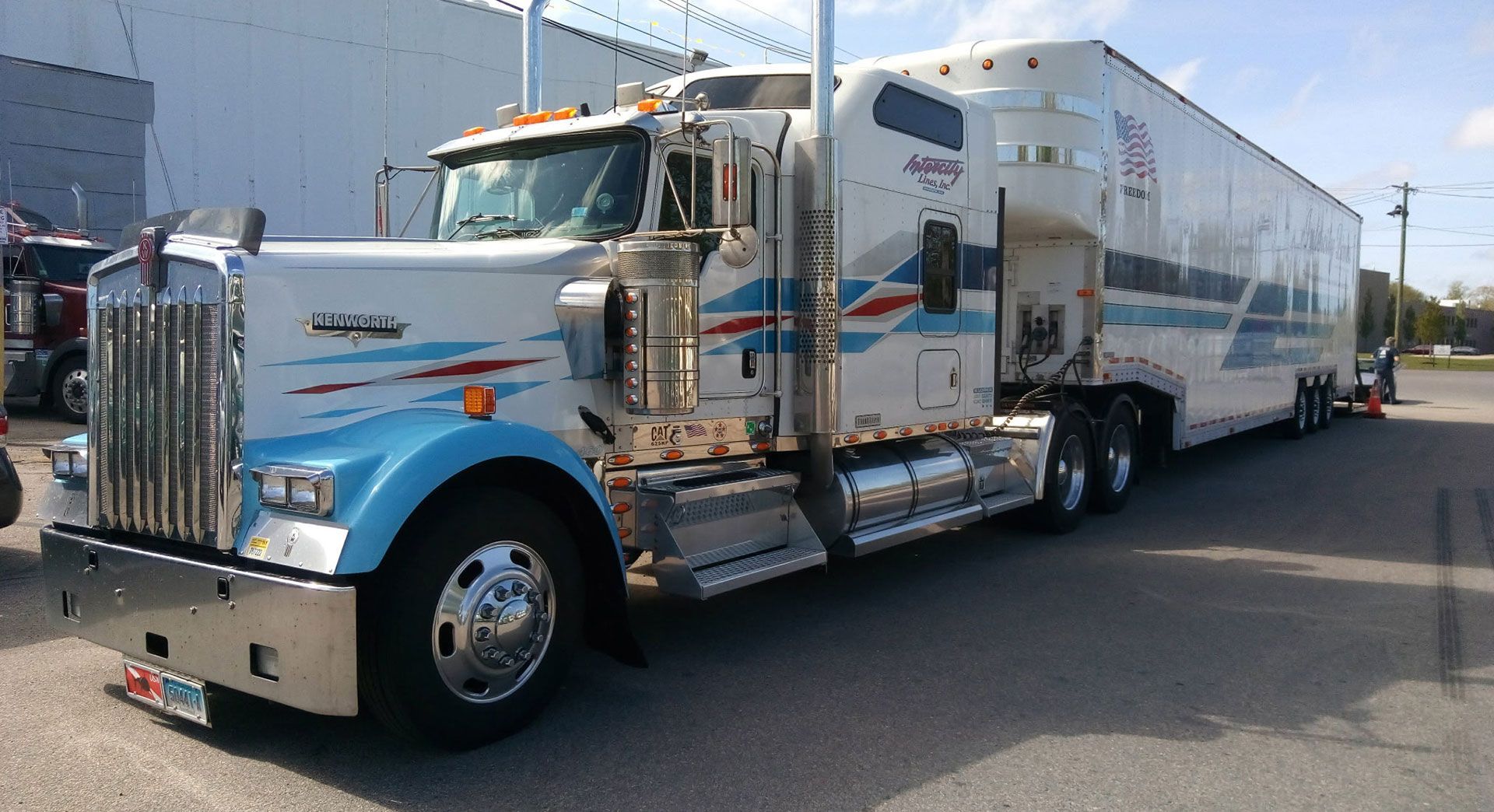 White semi-truck with a chrome grille pulling a long silver trailer on a sunny street, side view.