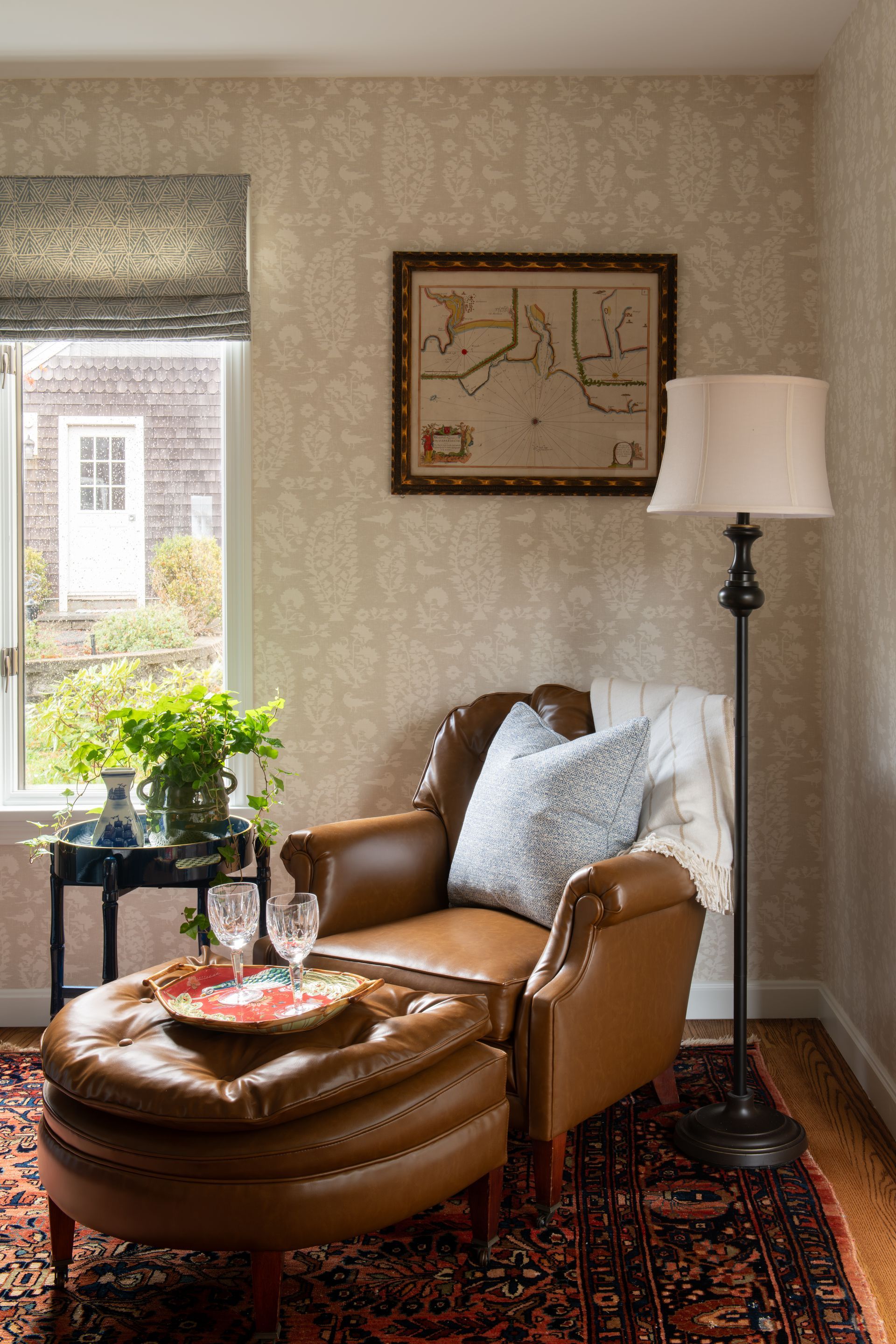 Cozy reading nook with leather chair, ottoman, floor lamp, and patterned rug. Window with blind.
