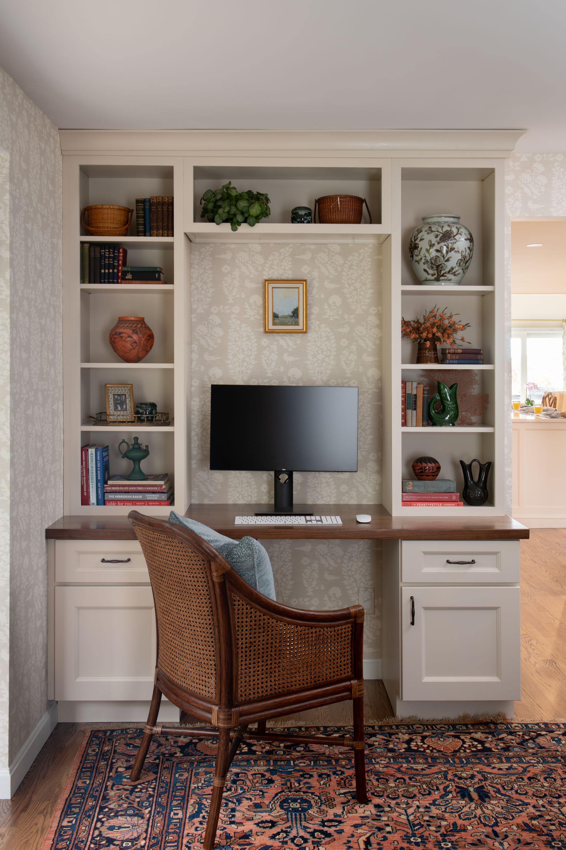 Built-in home office with desk and bookshelves. Wicker chair on patterned rug. Beige cabinets, wooden desk.