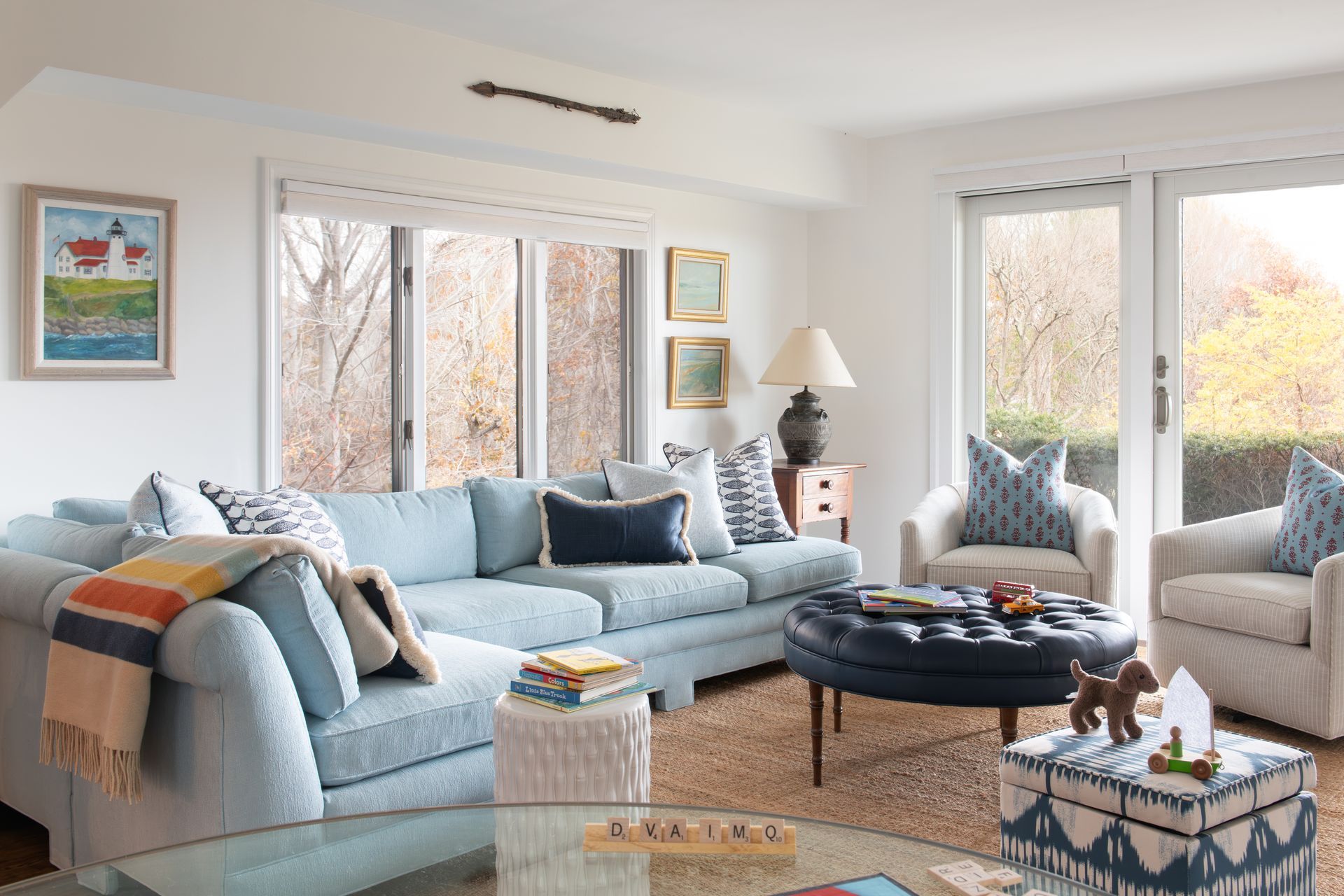 A light blue sectional sofa and armchairs in a bright living room, with a dark blue ottoman and outdoor views.