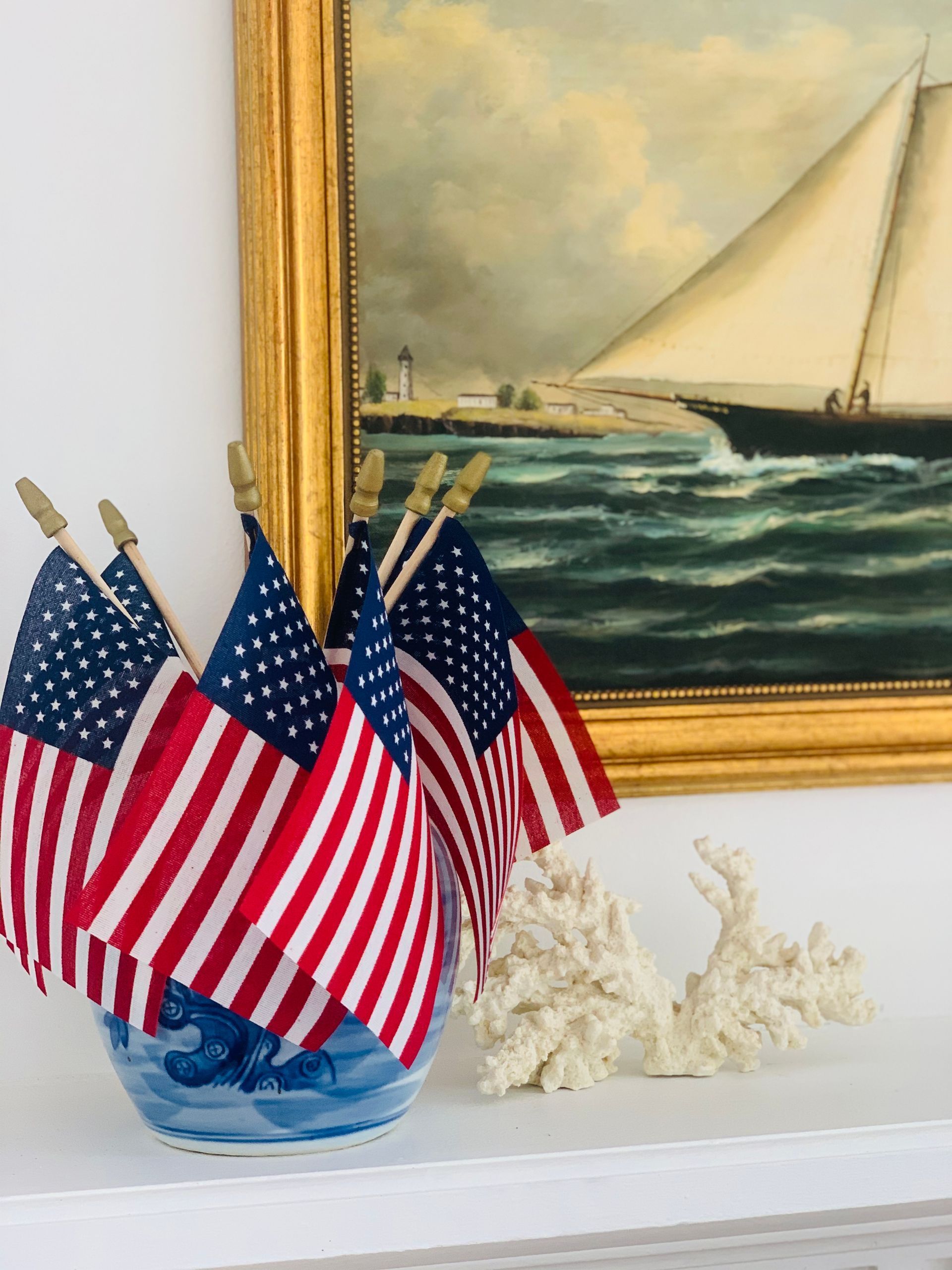 American flags in blue and white vase on a shelf, with a nautical painting in background.