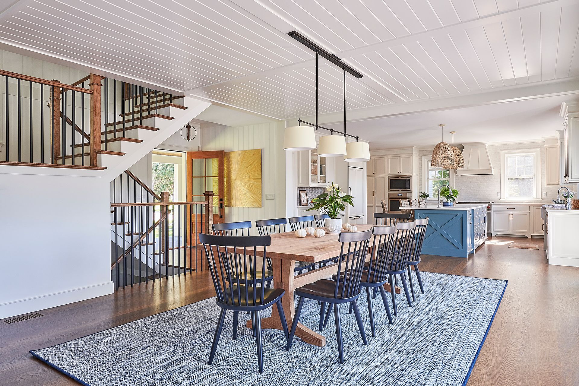 Dining room with wooden table, black chairs, rug, kitchen, and staircase.