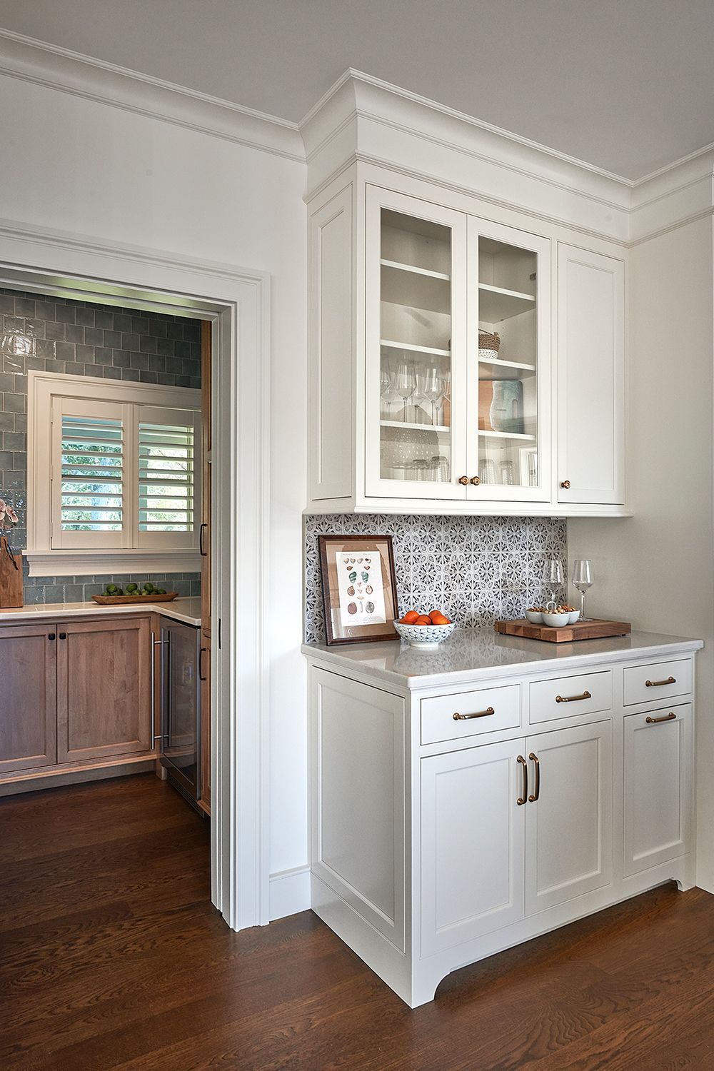 White built-in kitchen cabinet with glass-front upper cabinets, lower solid doors, and patterned backsplash, against a dark wood floor.