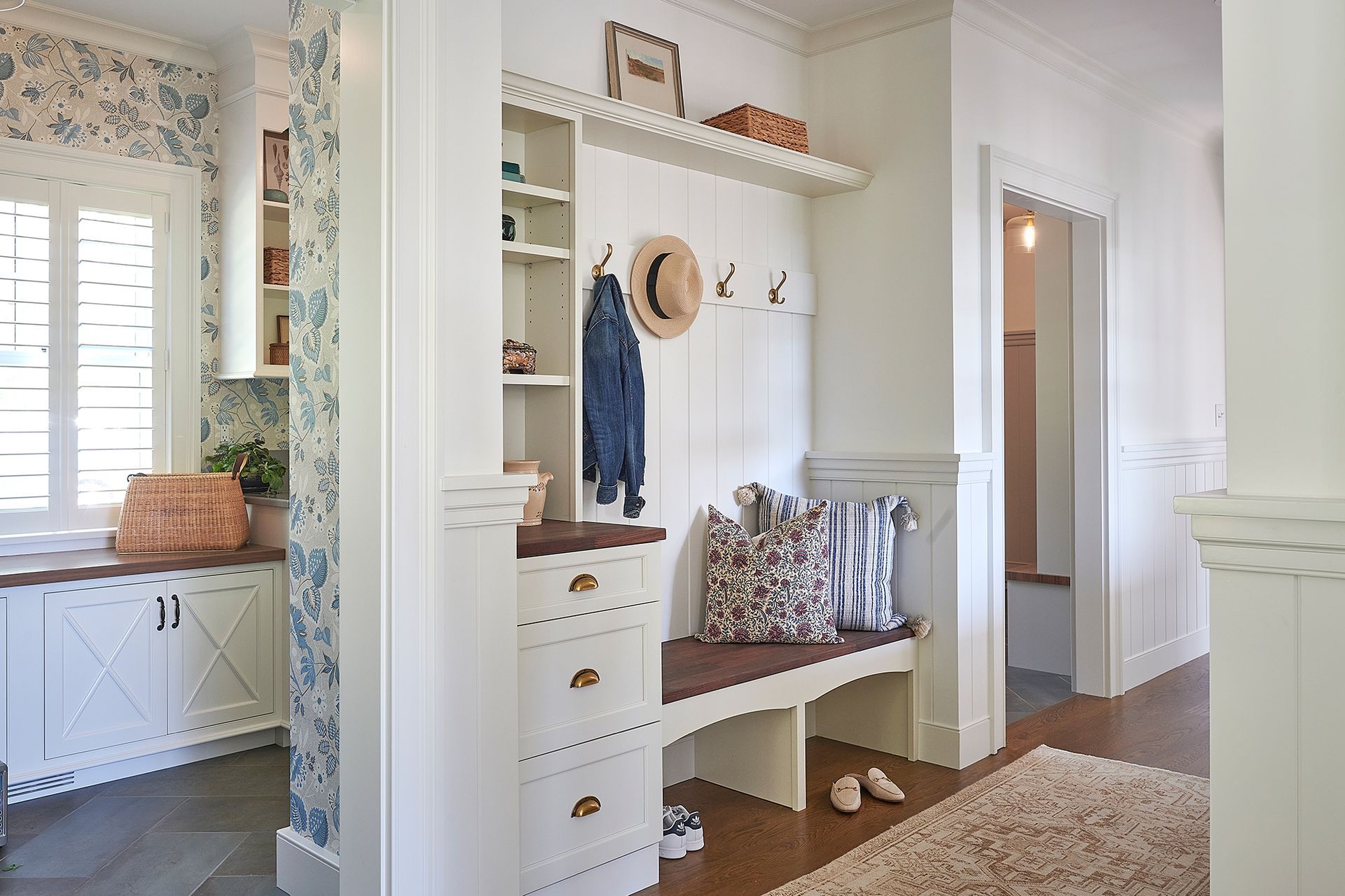 White entryway with built-in bench, shelves, and storage. Blue floral wallpaper, hat, jacket.
