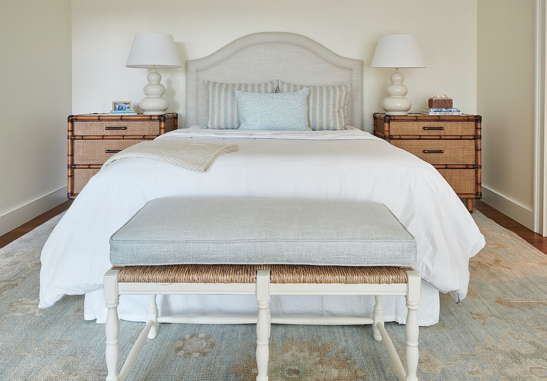 Bedroom with white bedding, rattan headboard, and bedside tables. A patterned bench sits at the foot of the bed.