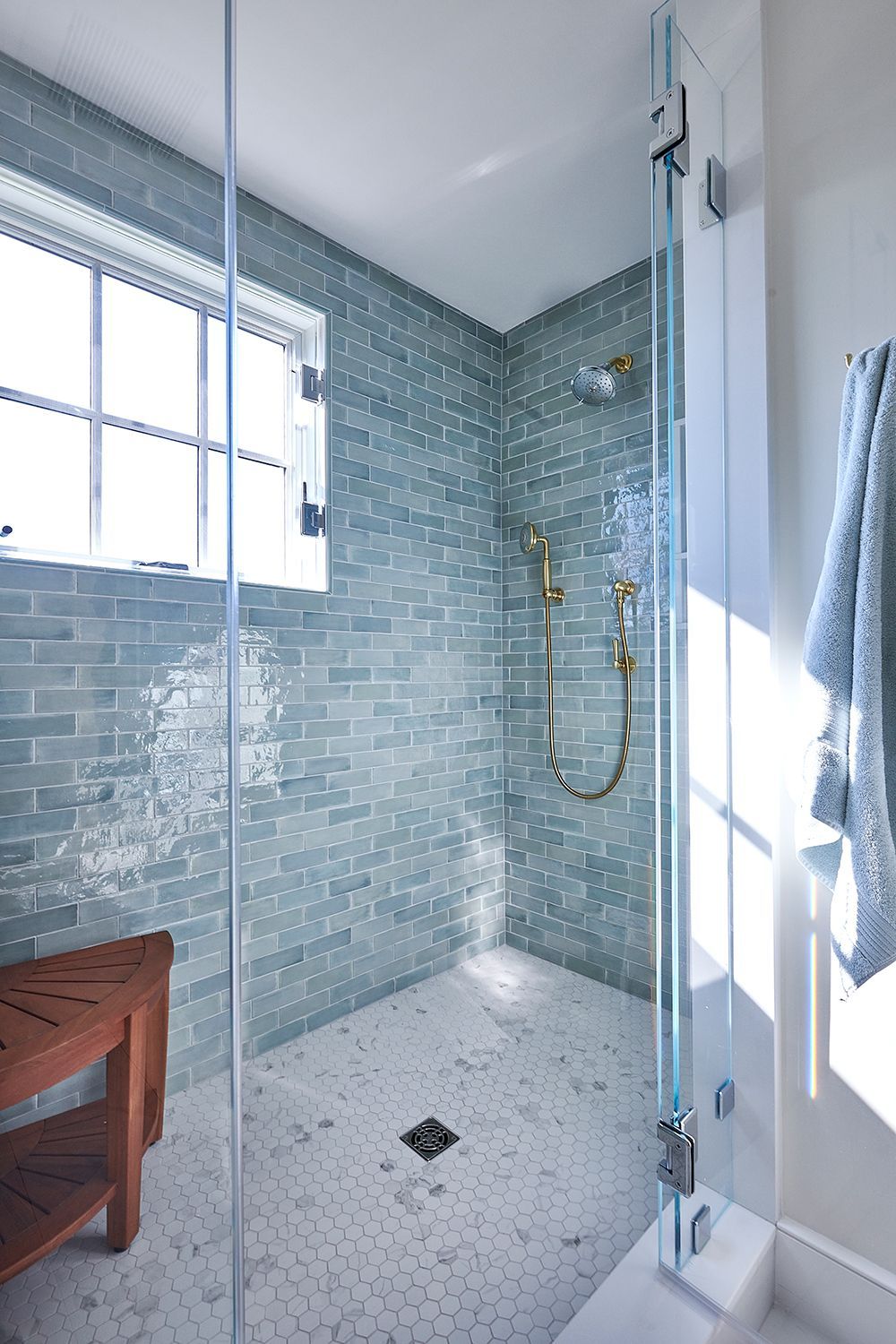 Shower with blue tiled walls, glass door, and gold fixtures. Wooden bench sits near a window.