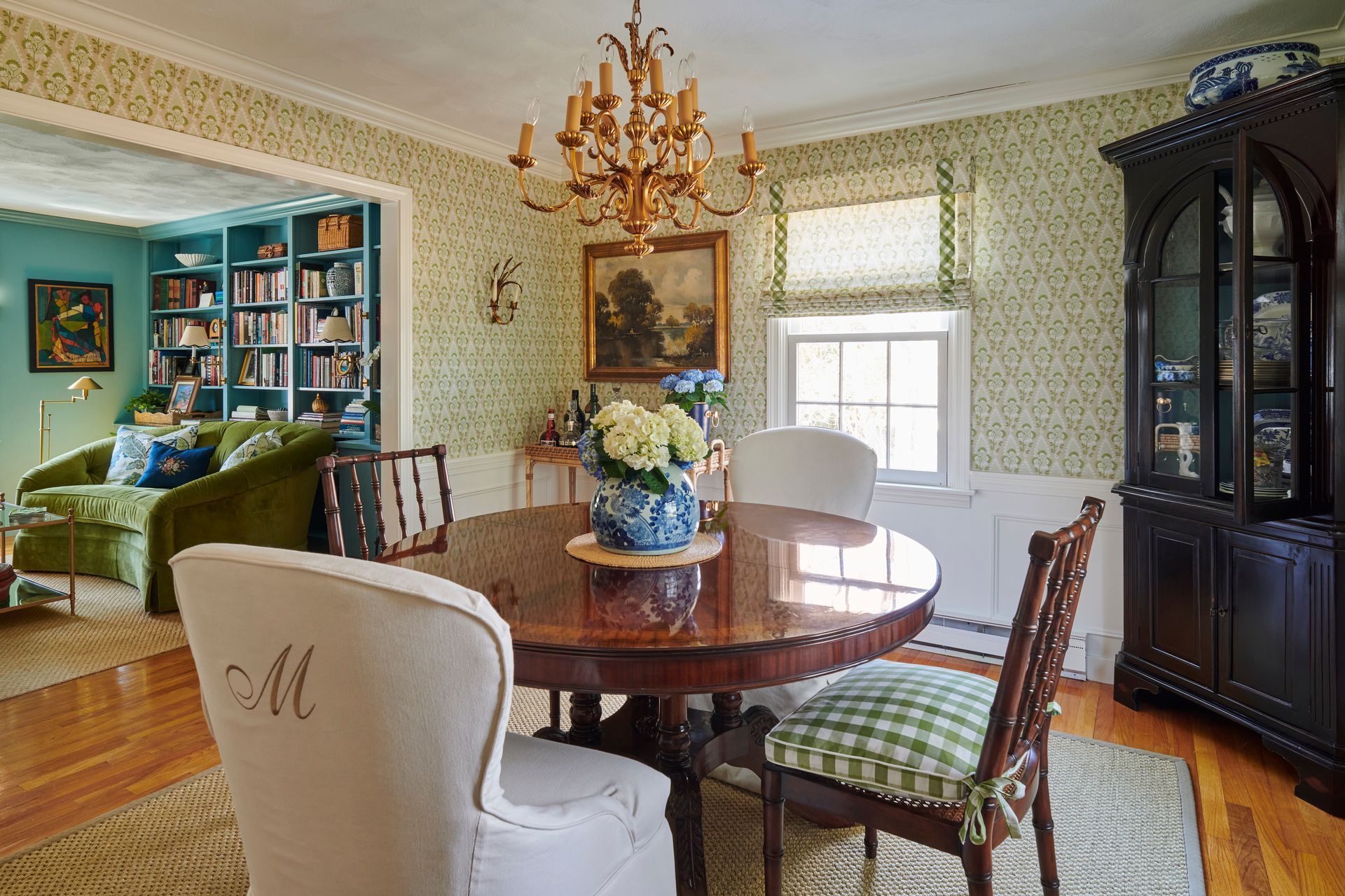 Dining room with round wooden table, chairs, chandelier, and built-in hutch. Green and white patterned wallpaper.