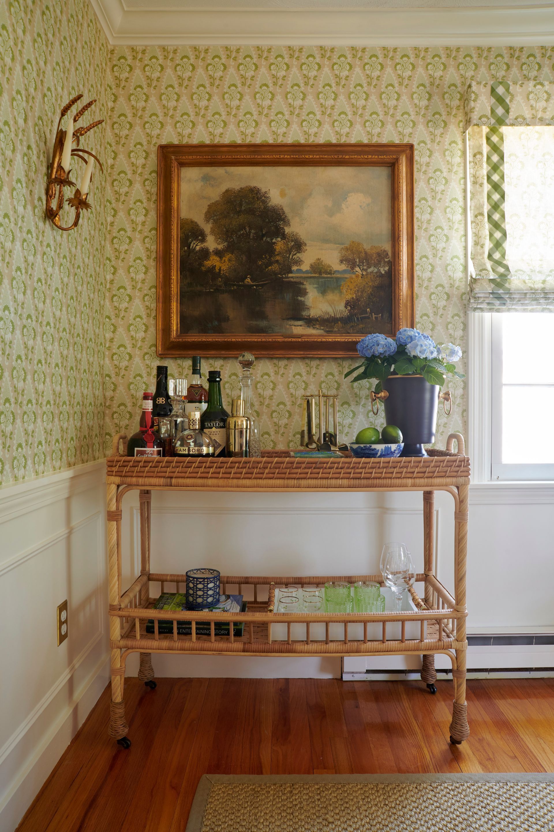 Rattan bar cart with bottles, painting, and blue flowers against floral wallpaper.