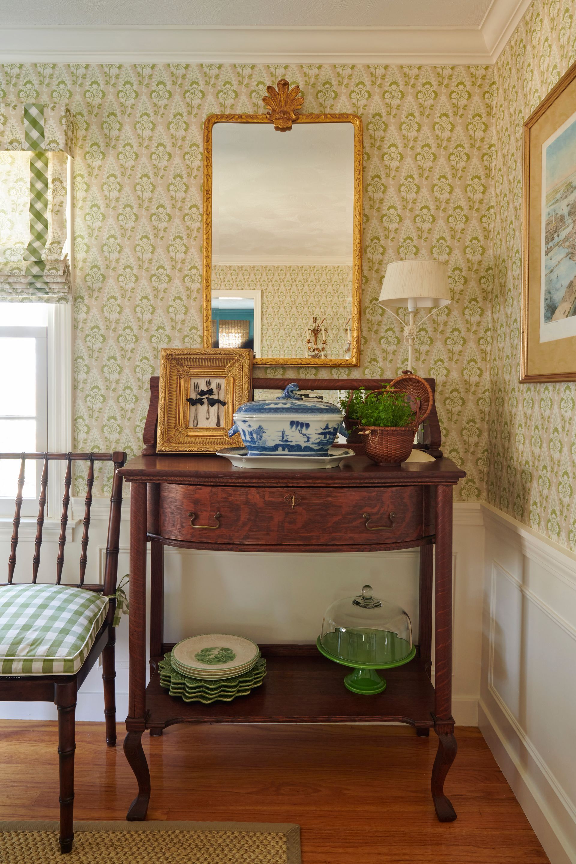A mahogany console table with decor, a gold-framed mirror, and floral wallpaper in a room with wood flooring.