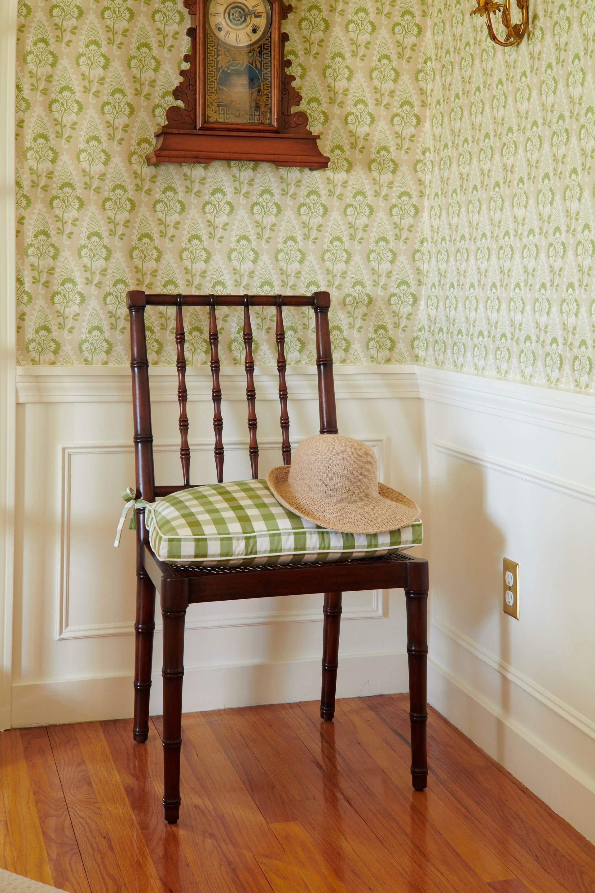 Chair with checkered cushion and straw hat beneath a wooden clock on wallpaper.