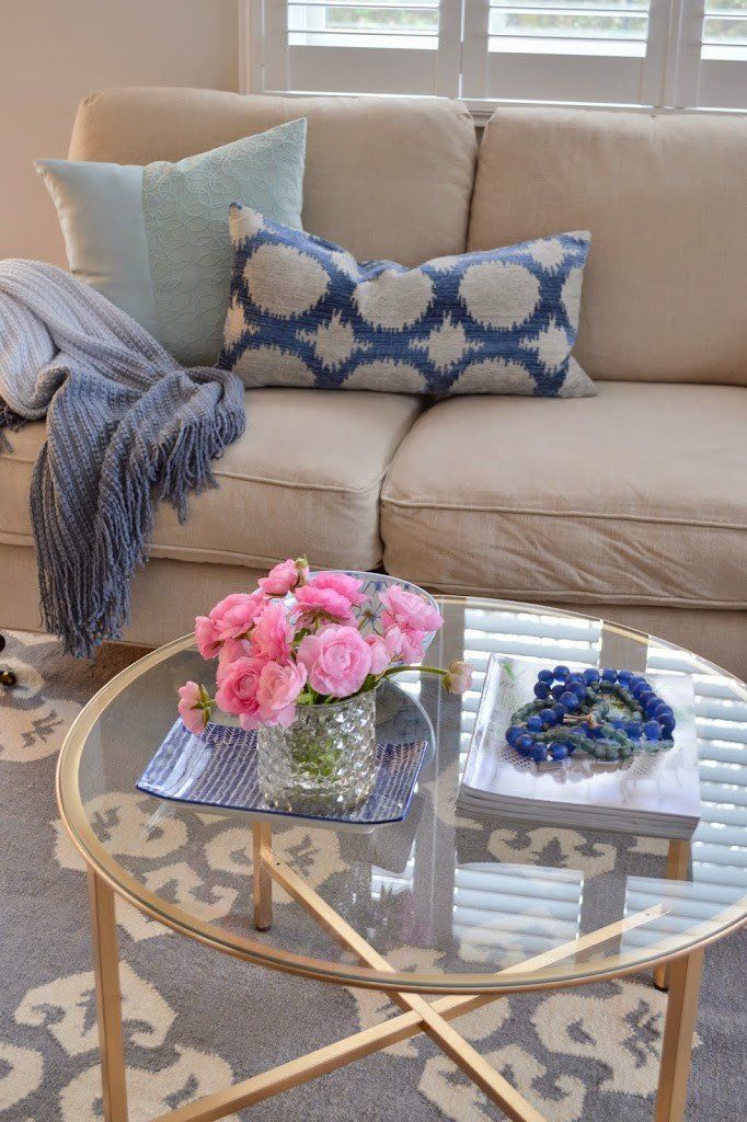 Beige couch with blue pillows and throw. Glass coffee table with flowers and decor.