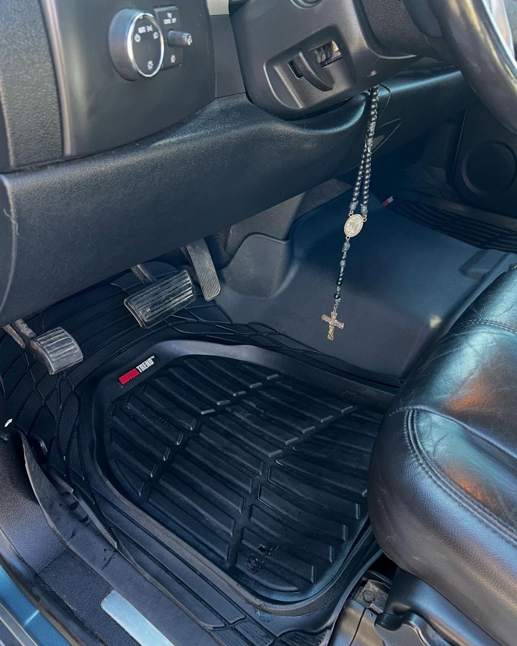 Black rubber floor mat in a car with pedals and a rosary hanging.