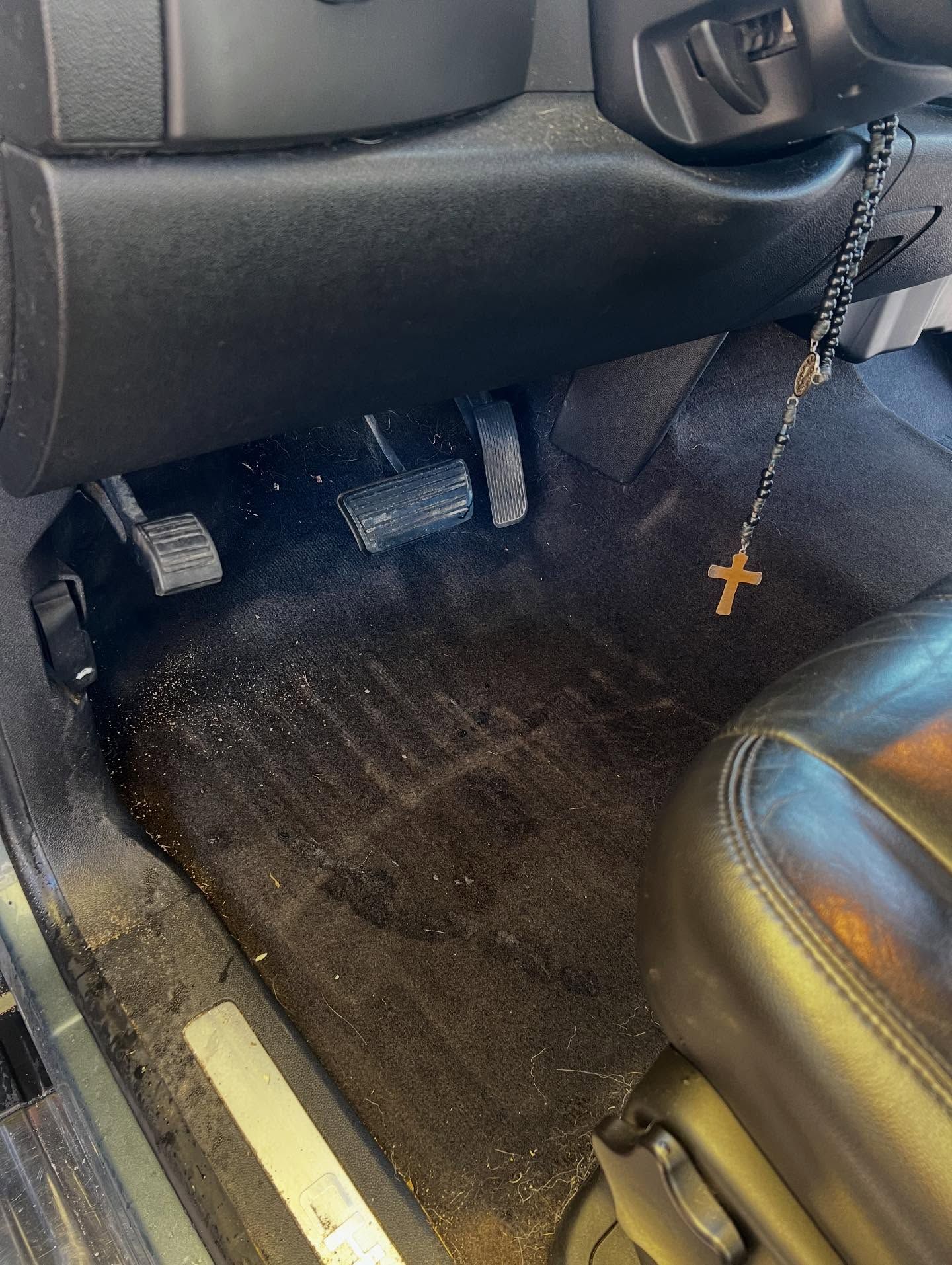 Car interior, dirty black floor mat with dust and debris, pedals, black seat, and rosary.