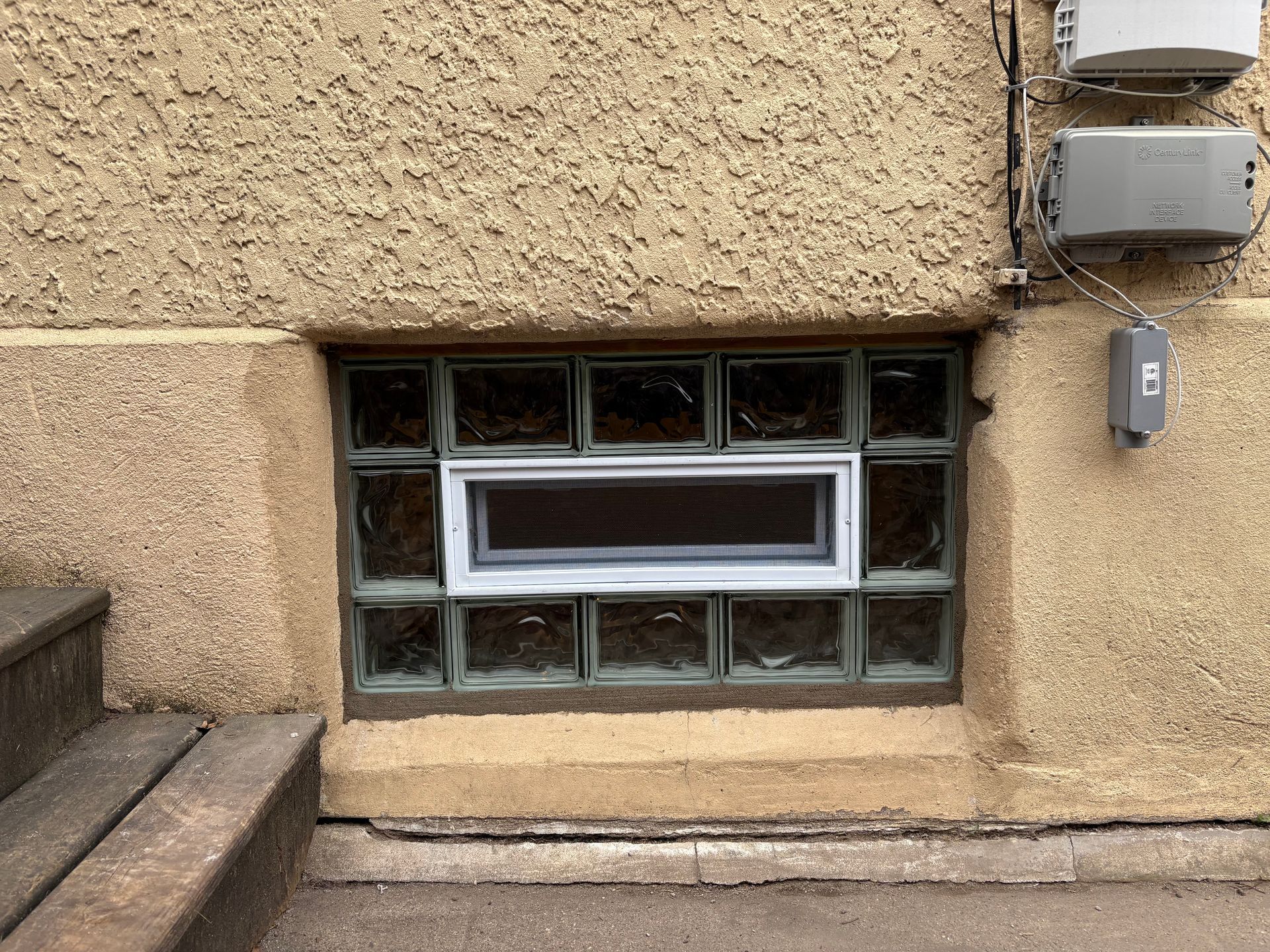 Basement window with glass block and white frame, below textured stucco wall and electrical meters.