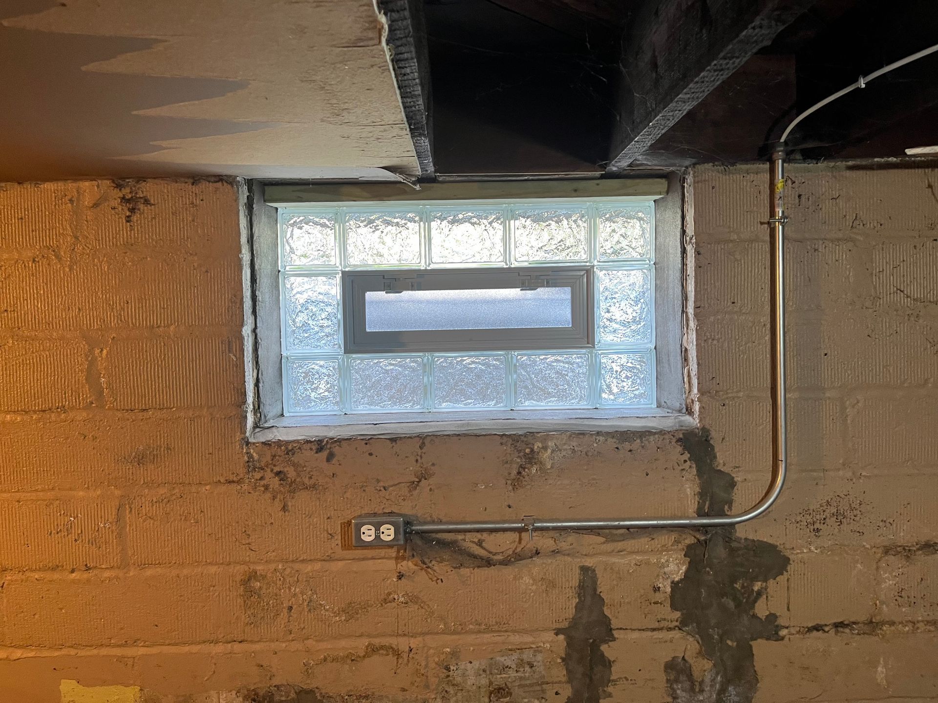 Basement window with glass blocks, electrical outlet and conduit. Concrete walls show water stains and aging.